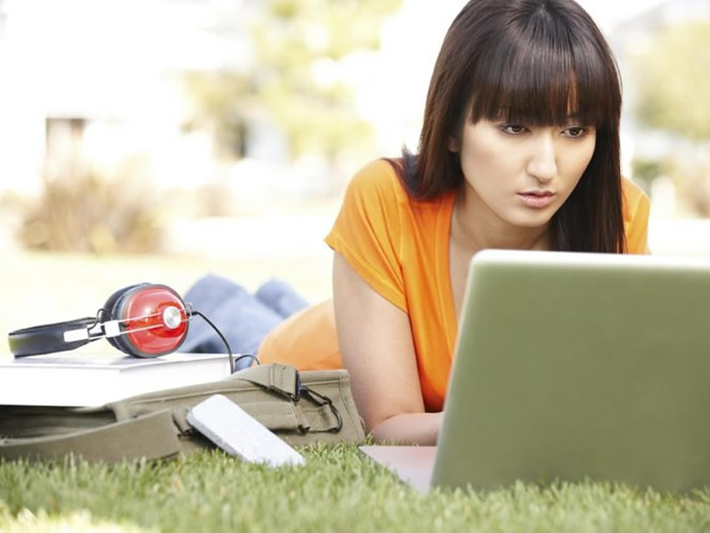 Young woman lying on the grass, researching college financial aid on her laptop