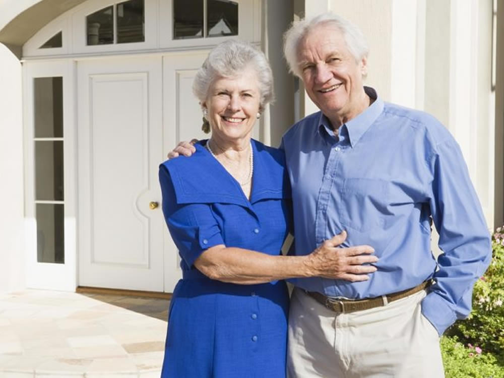 Smiling older couple in front of house they financed with reverse mortgages in retirement