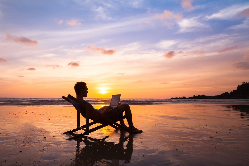 Man sitting on beach with laptop has mastered the art of knowing how to simplify your finances